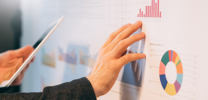 Person analyzing data on a large digital display screen featuring bar charts, pie charts, and graphs, while holding a tablet — representing business analytics, data visualization, and tech-driven decision making