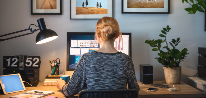 Person working at a modern desk setup with a computer, digital clock, lamp, tablet, notebooks, and framed wall art, ideal home office or study environment