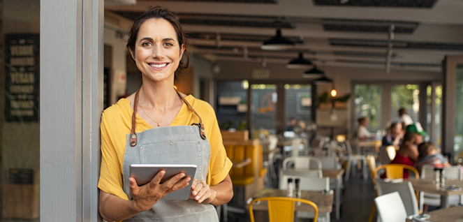 Restaurant staff member in a yellow shirt and grey apron holding a tablet, standing in a modern café with tables, chairs, and natural light from large windows — representing hospitality, customer service, and digital ordering systems.