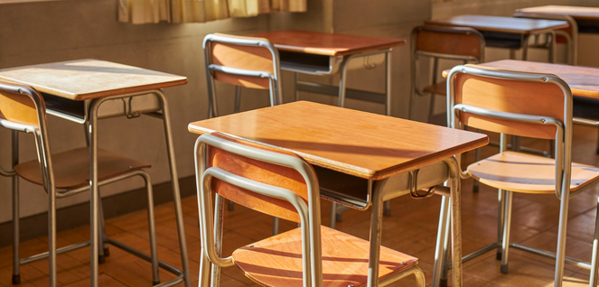 Empty classroom with wooden desks and metal-framed chairs arranged in rows on a polished wooden floor, illuminated by natural sunlight streaming through large windows — representing education, learning spaces, and academic environments.