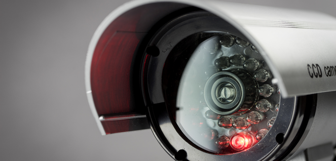Close-up of a CCD security camera with visible lens and infrared LEDs, one glowing red, housed in a metallic casing with a protective cover — representing surveillance technology, property security, and monitoring systems.