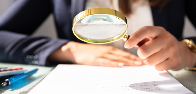 erson holding a magnifying glass over a document on a desk, suggesting close examination or review.