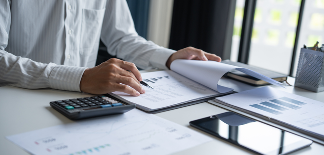 Person working at a desk with documents, charts, graphs, a calculator, and a tablet, indicating financial analysis or data review.