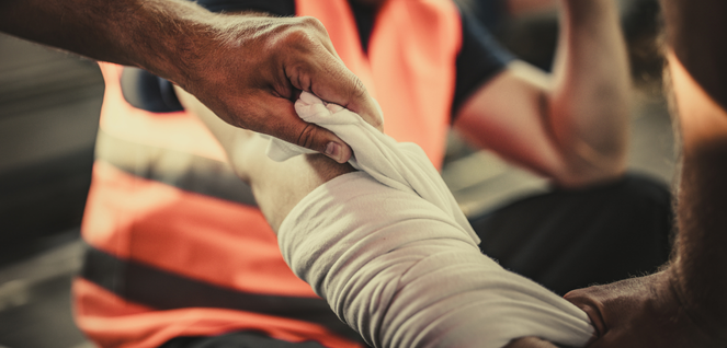 Close-up of hands wrapping a bandage around an arm, with one person wearing an orange safety vest — representing first aid, emergency response, workplace safety, and injury care.