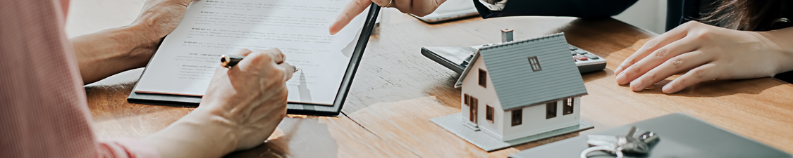 Close-up of hands reviewing and signing a property contract at a table, with a model house, keys, and calculator, symbolising legal review and agreement in a private real estate transaction.