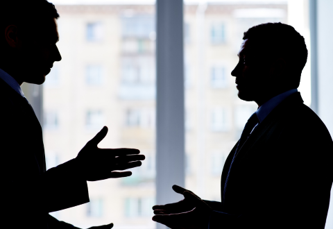 Silhouettes of two professionally dressed men engaged in a conversation in front of large office windows. Their hand gestures suggest active discussion, with a softly blurred urban building visible in the background, emphasizing themes of negotiation, communication, or conflict resolution.