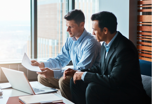 Two professionally dressed men seated in a modern office space, reviewing a document together near a large window with a cityscape view. One man gestures toward the paper while the other listens attentively, suggesting a collaborative business discussion.