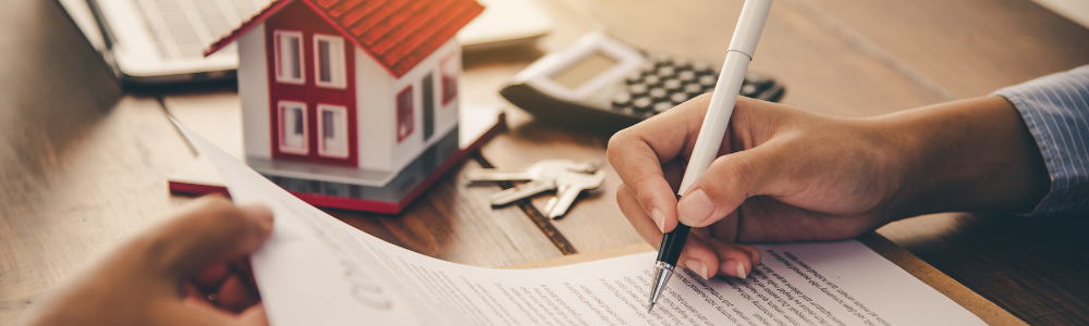 Close-up of a person signing a property contract at a wooden table, with a model house, keys, and calculator, symbolising legal review and financial preparation before bidding at auction.